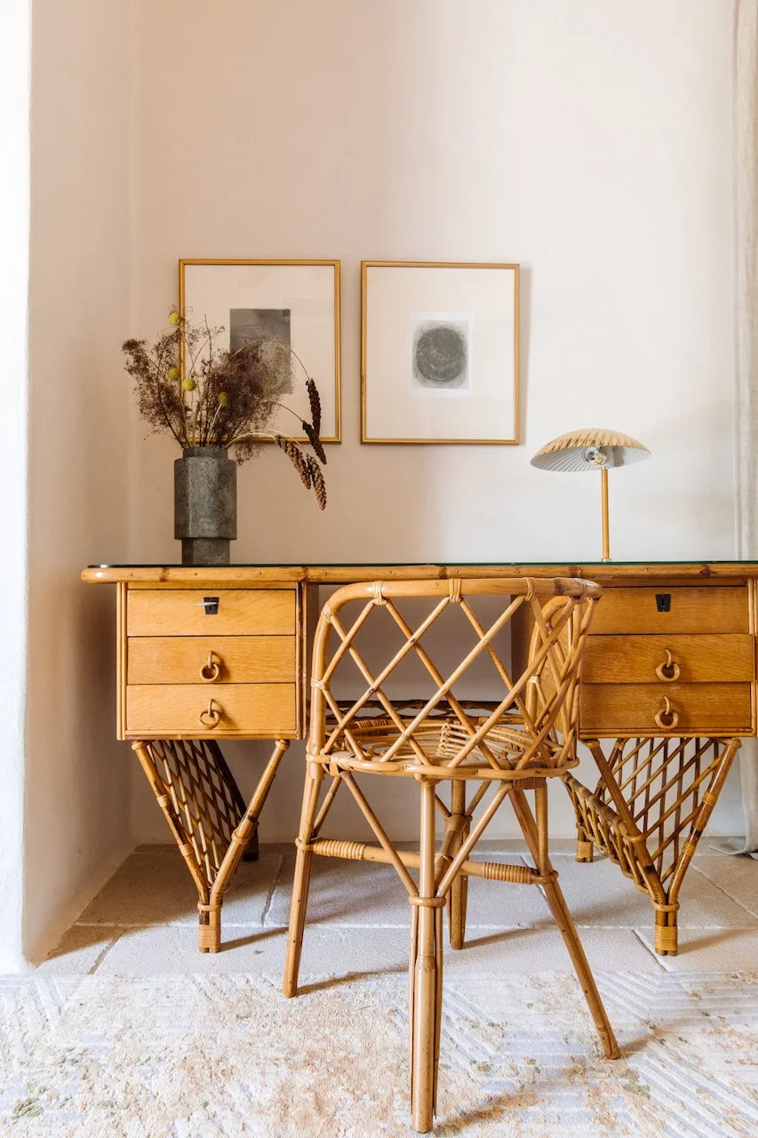 A vintage wooden desk with rattan legs and a matching rattan chair in front of a white wall. On top of the desk is a vase with dried flowers and a small, modern table lamp with a shell-shaped shade. Two framed abstract art prints hang on the wall abo