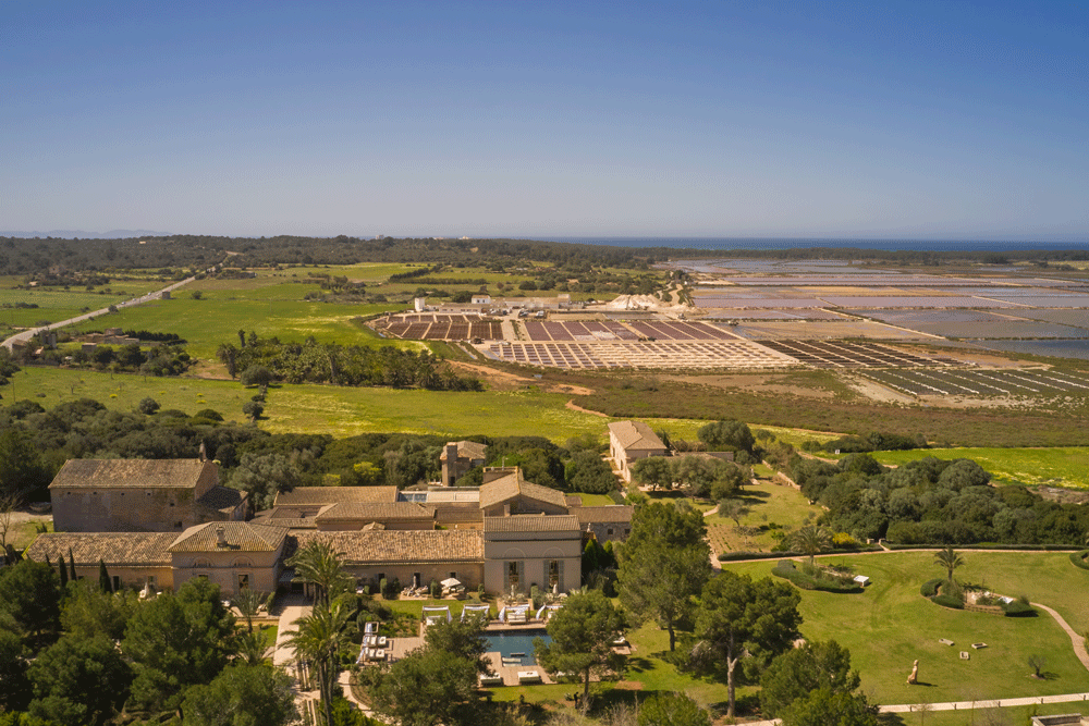 Aerial view of a large estate with a mansion, swimming pool, lush green lawn, and surrounding trees, with salt flats or agricultural fields in the distance under a clear blue sky.