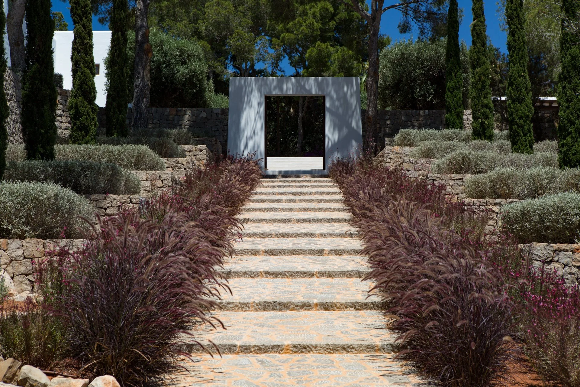 Stone steps leading up to a white modern archway surrounded by greenery and tall trees in a landscaped garden.