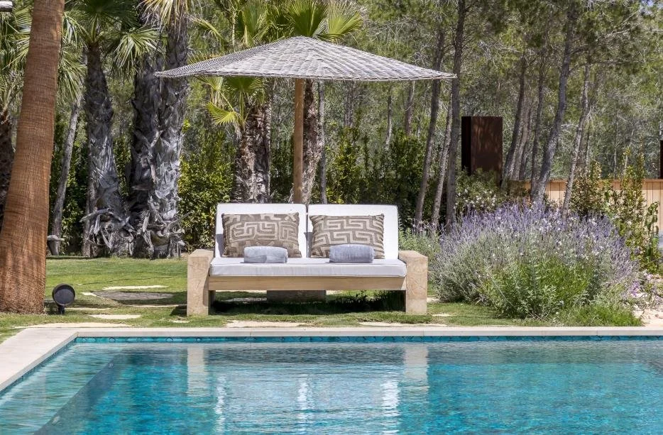 A backyard scene with a swimming pool, a white cushioned bench with two pillows, an umbrella, lush grass, and tall trees and bushes in the background.