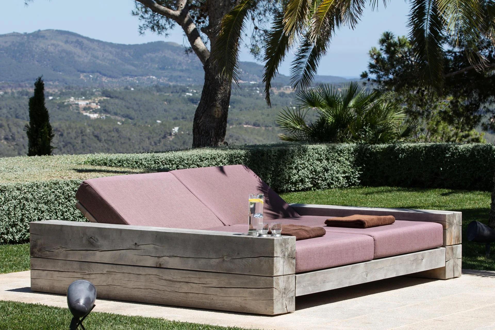 Outdoor patio with a wooden couch with pink cushions, a glass of water, and brown towels. Background features lush green trees, a hedge, and distant hills under a clear blue sky.
