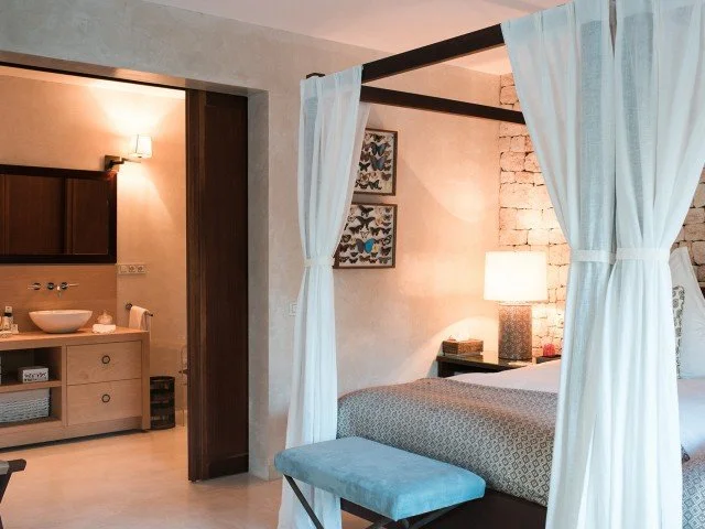 Bedroom with a four-poster bed with white curtains, a blue bench at the foot, and a lit bedside lamp. An exposed brick wall is behind the bed, and a small framed artwork hangs above.