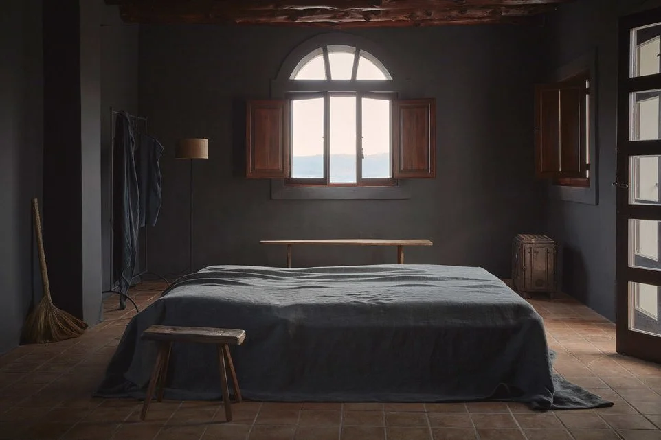 A bedroom with dark gray walls, a wooden bed frame with a dark gray bedspread, a small wooden bench at the foot of the bed, an open window with wooden shutters, a floor lamp, and minimal decor.