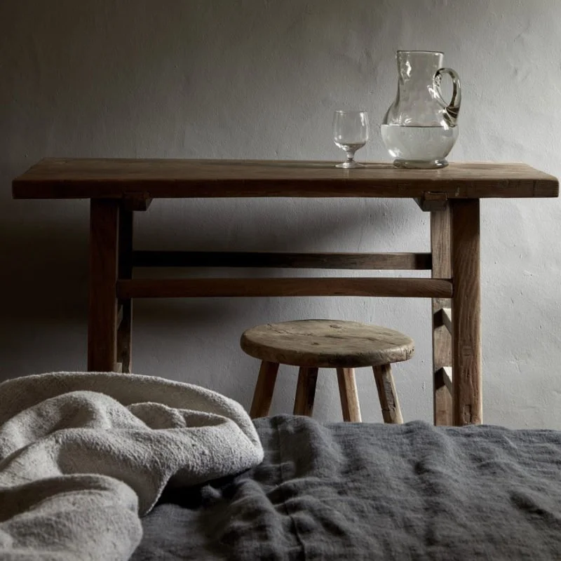A rustic wooden table with a glass pitcher and a glass of water on top, a small wooden stool below, and a bed with a blanket in the foreground.