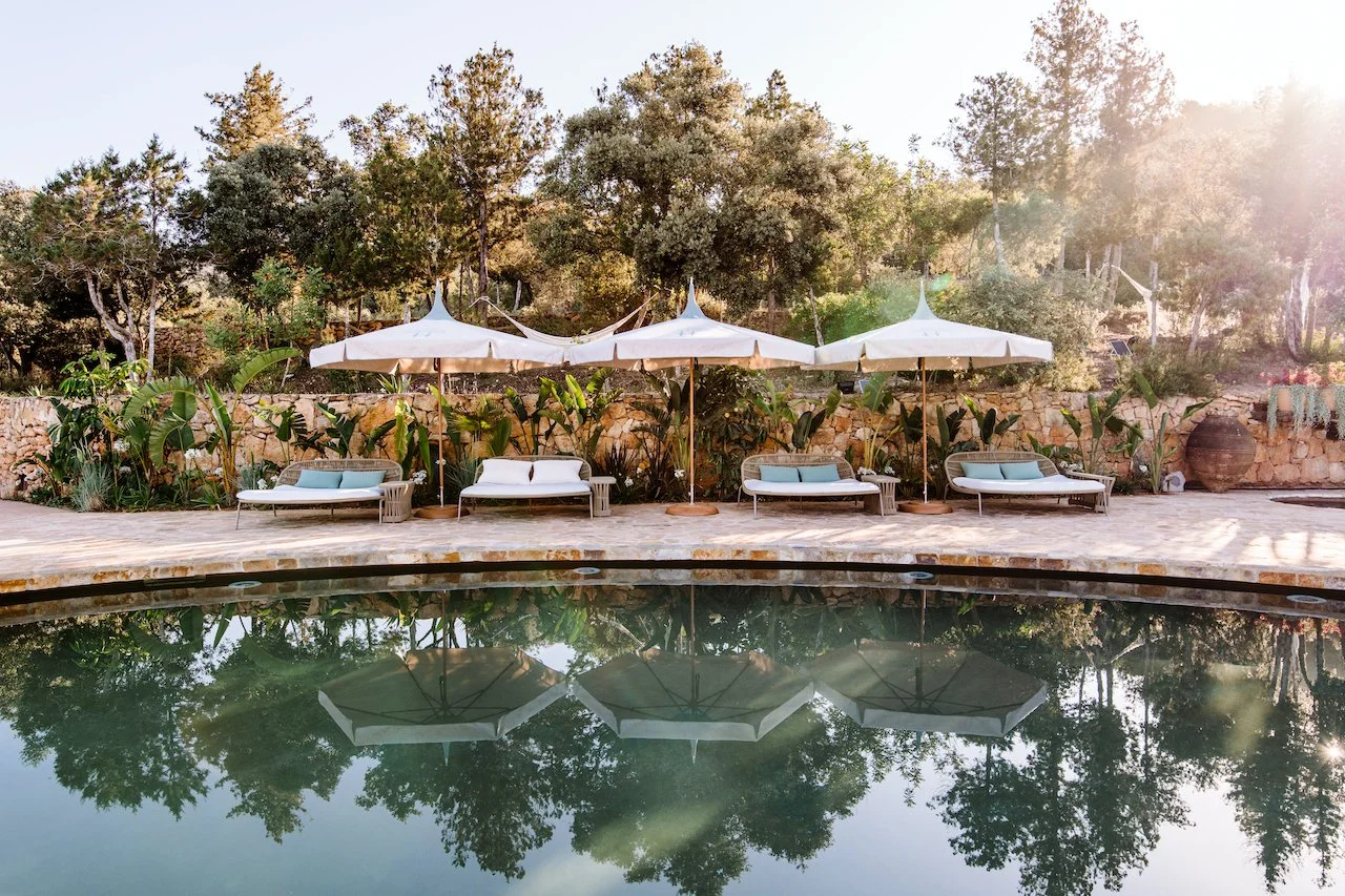 Poolside scene with four lounge chairs, each with a pillow, and four large white umbrellas, set against a background of lush trees and a stone wall, with the reflection of the scene visible in the pool water.