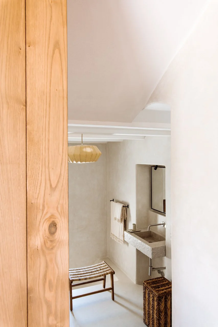 Interior of a minimalist bathroom with a small stone sink, a mirror, a towel hanging on a black rod, a wooden stool, a woven basket, and a hanging beige pendant light.