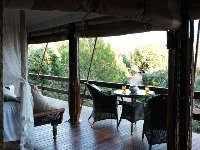 Outdoor patio with a round table set with four glasses and a small bowl, surrounded by four black wicker chairs, under a canopy with a view of green trees and bushes.