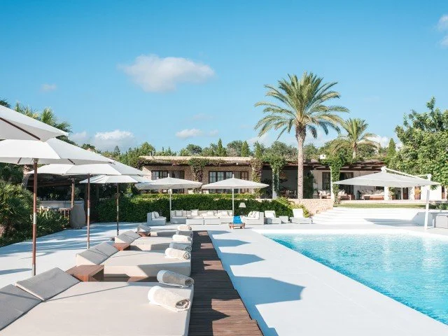 Luxury poolside area with lounge chairs, umbrellas, and a large swimming pool, surrounded by palm trees and modern buildings under a blue sky.