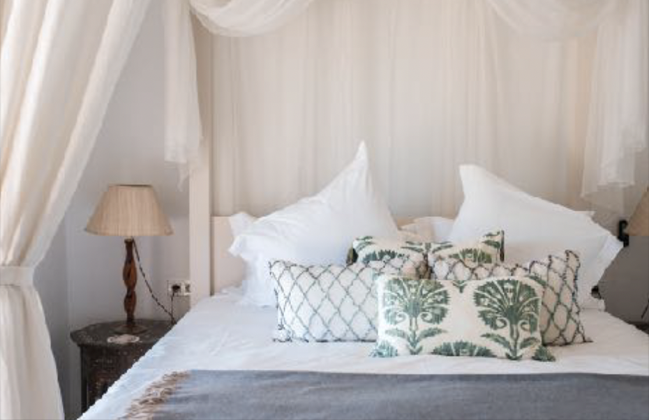 A neatly made bed with white linens and multiple pillows, including two large white pillows and several decorative pillows with green and white patterns. There are bedside lamps with beige shades on both sides of the bed.