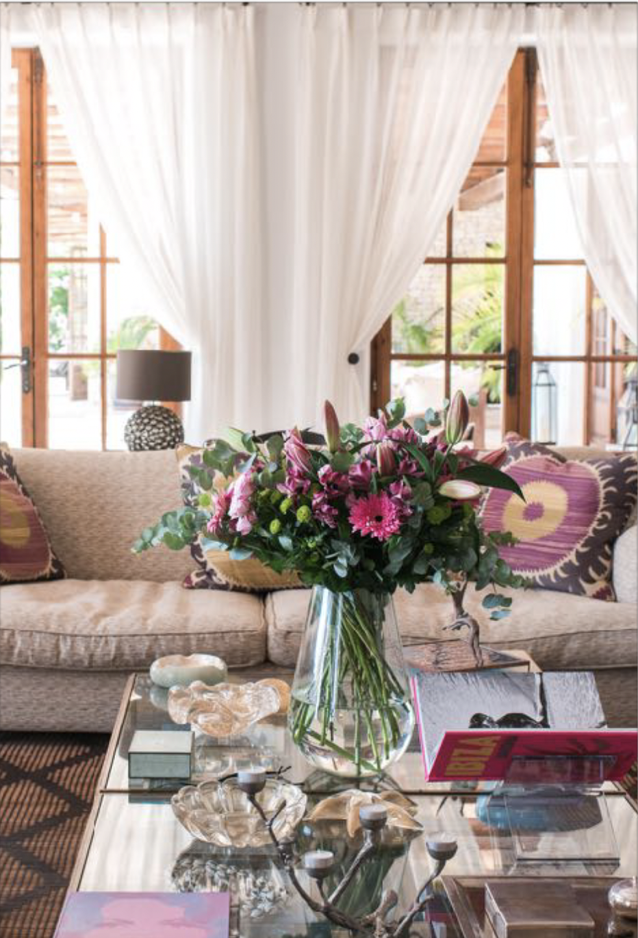 Living room with a beige sofa, decorative pillows, glass coffee table with jewelry and a pink magazine, a large bouquet of pink lilies and flowers on the table, wooden-framed windows with white curtains, and a black table lamp in the background.