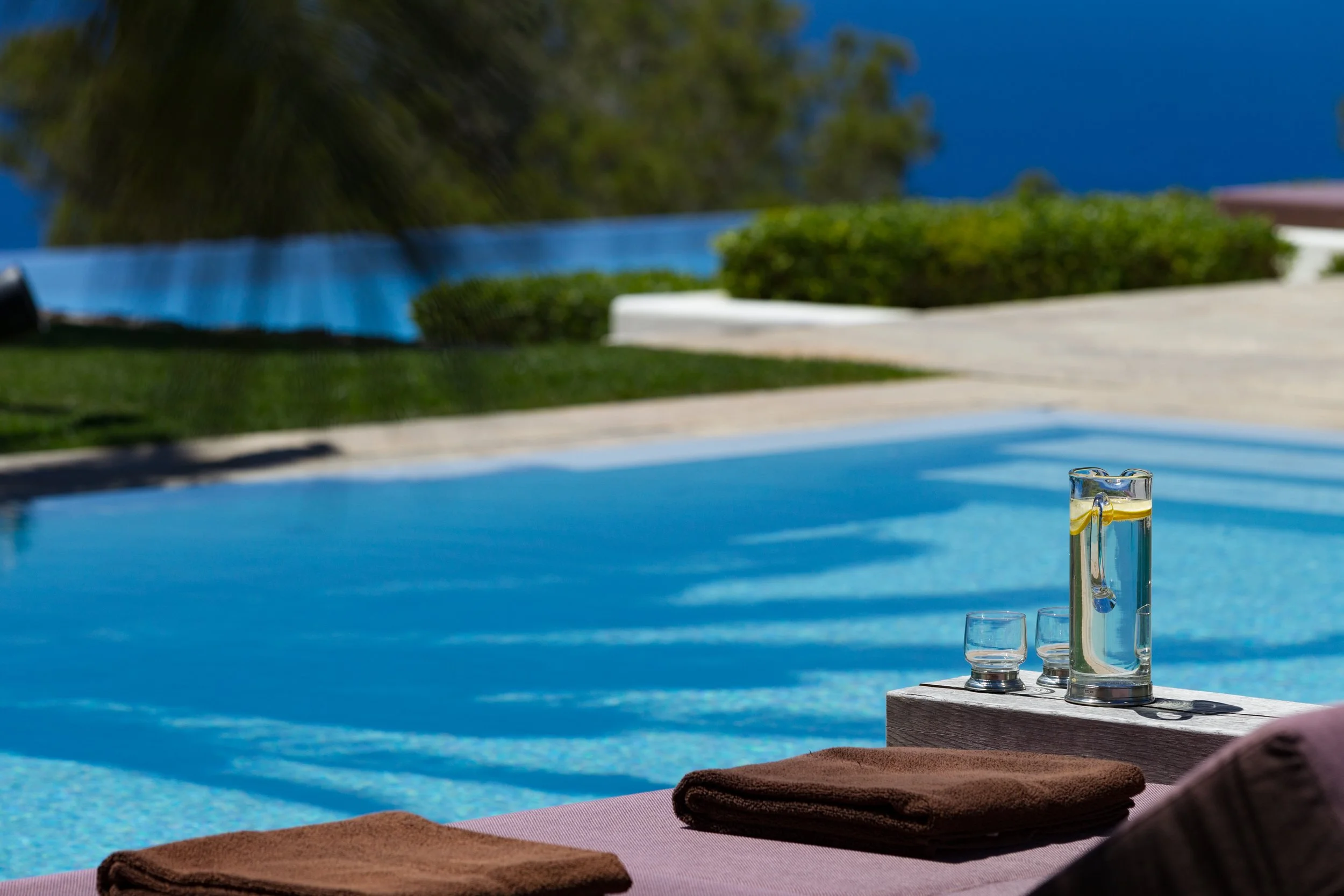 A poolside scene with a wooden table holding a set of glassware, including a decanter and glasses, near a lounge chair. In the background, the pool and a garden with bushes and trees are visible under a bright blue sky.