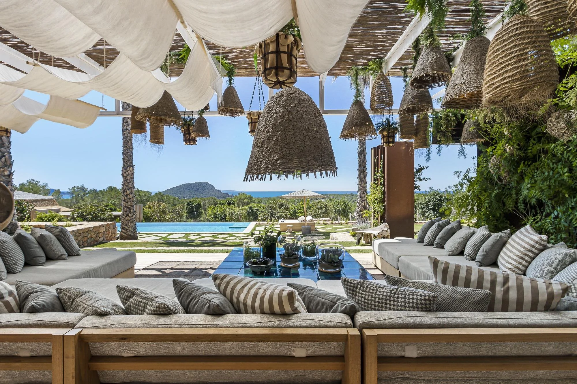 Outdoor lounge area with large beige sectional sofas, striped throw pillows, glass-topped coffee table, and a view of a swimming pool, trees, and hills in the distance. Hanging wicker and fabric pendant lights are suspended from a pergola roof, with 