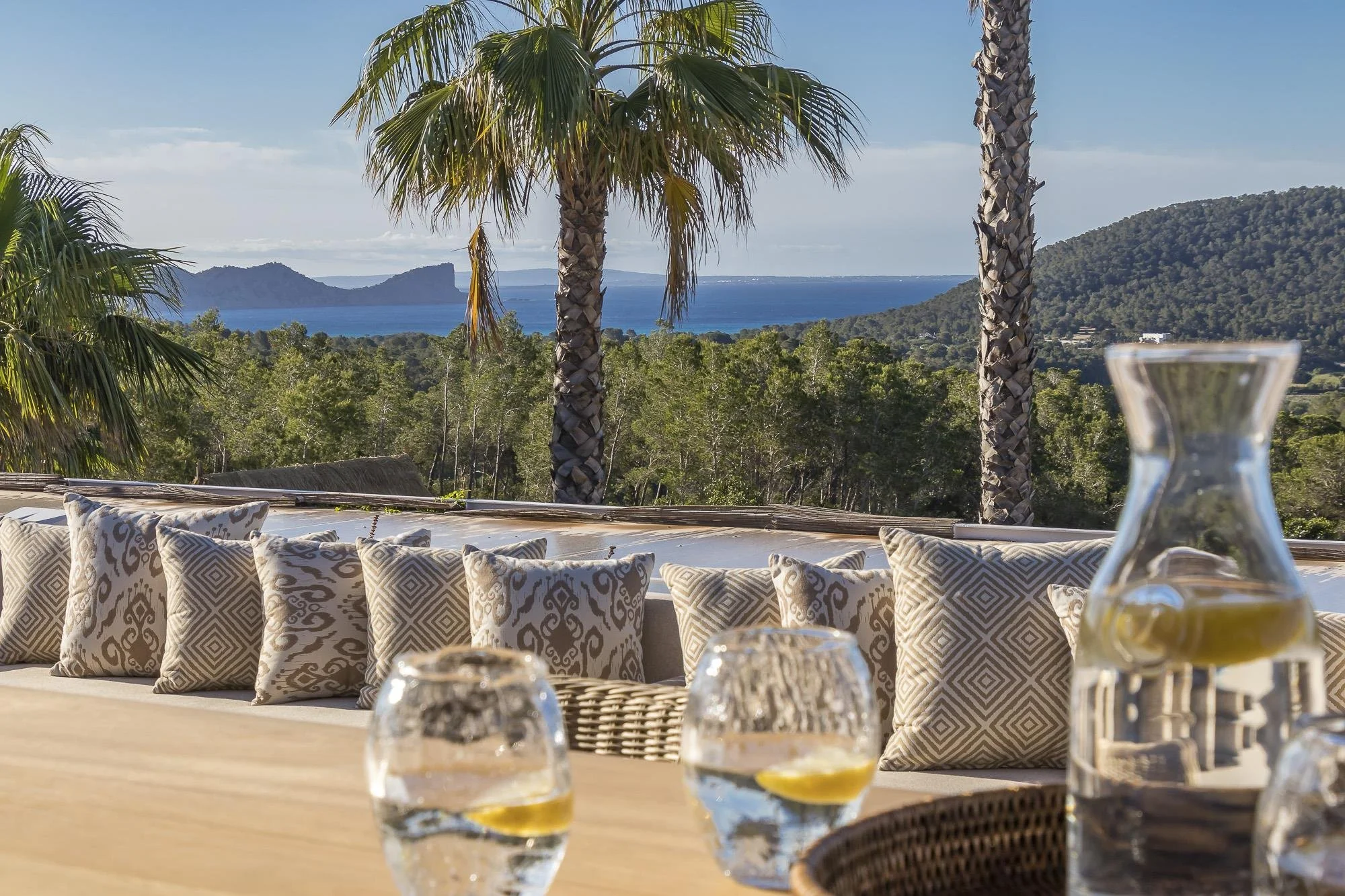 Outdoor patio with patterned cushions, palm trees, lush green forest, distant mountains, and ocean view in the background. Glasses with water and lemon slices on the table.