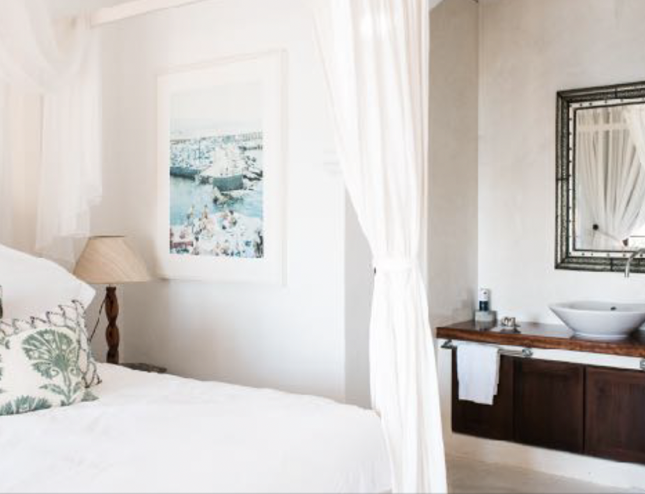 A bedroom with a white bed, a bedside lamp, framed artwork on the wall, and an open closet with a curtain. There is a bathroom sink with a mirror and a towel on a wooden vanity.