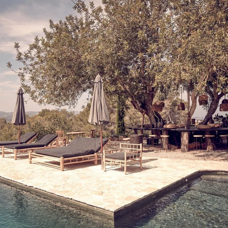 A poolside lounge area with black cushioned sun loungers, gray umbrellas, and outdoor furniture under a large tree with hanging baskets, on a sunny day.