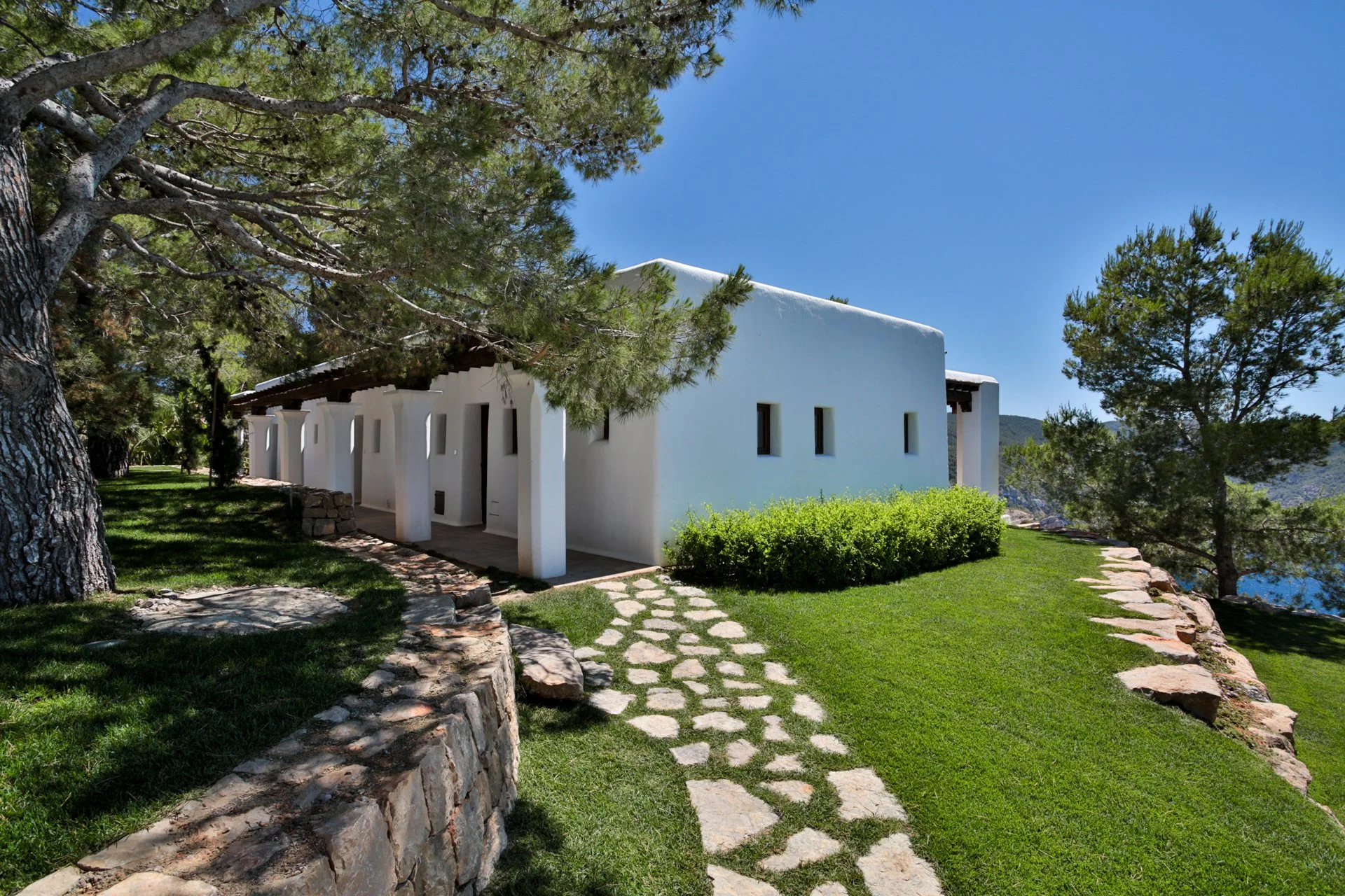 A white modern house with a flat roof and small rectangular windows, surrounded by green grass and trees, with a stone pathway leading to the entrance, under a bright blue sky.