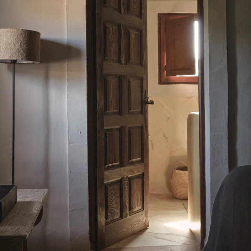 Indoor scene showing a partially open wooden door leading to a room with a window, a basket, and a pedestal.