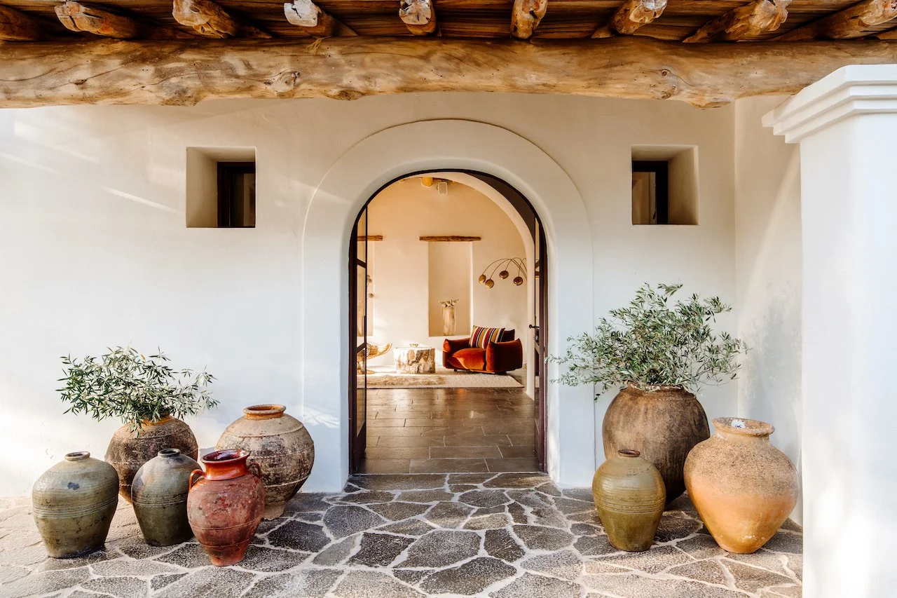 Entrance to a Mediterranean-style home with white stucco walls, a stone floor, and large rustic clay pots with plants near the doorway, leading to a cozy interior with a red velvet chair and modern decor.