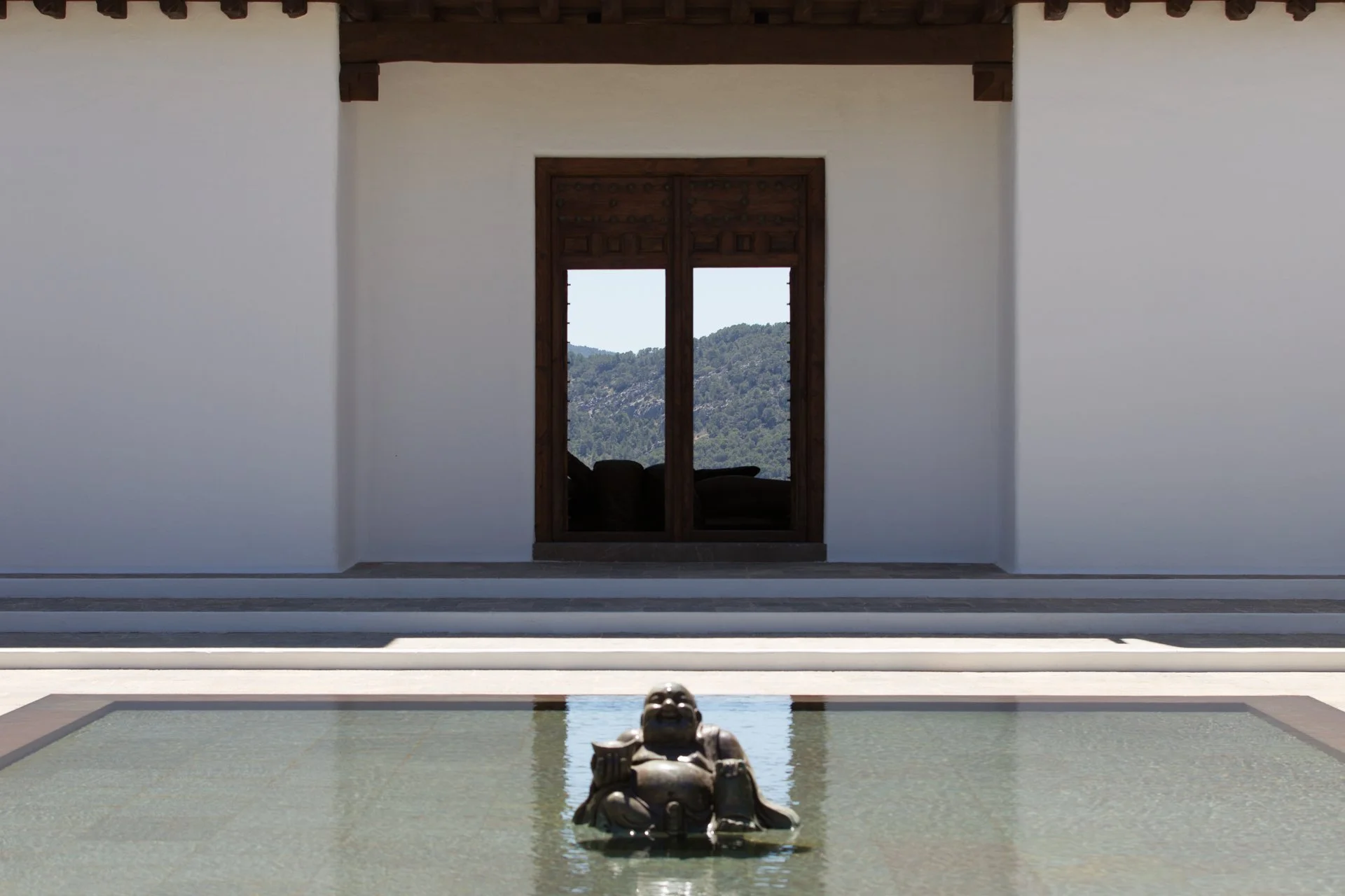 A courtyard with a rectangular water feature and a sculpture of a reclining figure in the foreground. In the background, there's a large open window with a view of a mountain landscape, flanked by white walls and a wooden overhang.