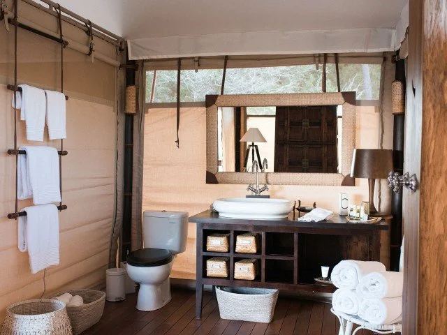 Bathroom with wooden walls, a toilet, a mirror above a dark wood vanity with a white vessel sink, a table lamp, and rolled towels.