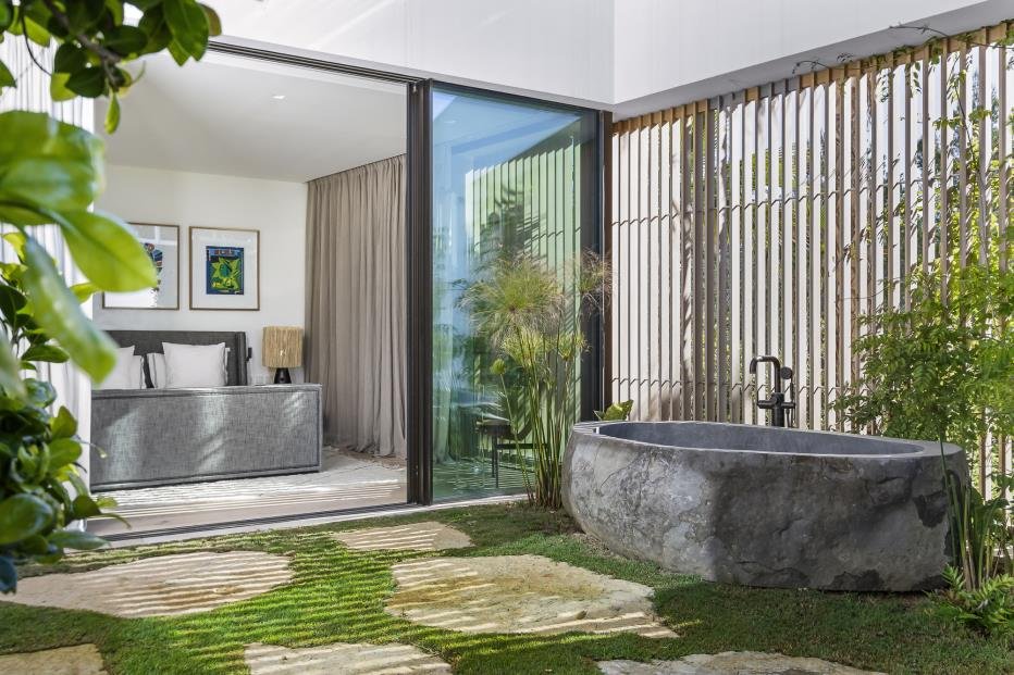 Modern bedroom visible through sliding glass doors, with a large bed, artwork on the wall, and natural light. Outdoor patio area with a stone bathtub, lush green plants, and a wooden privacy screen.