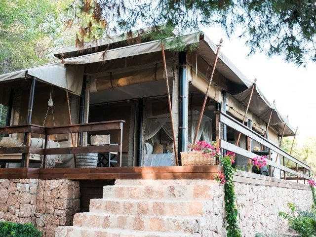 Outdoor view of a rustic porch with brick steps, wooden railing, and a canvas-covered roof, surrounded by trees and flowers.