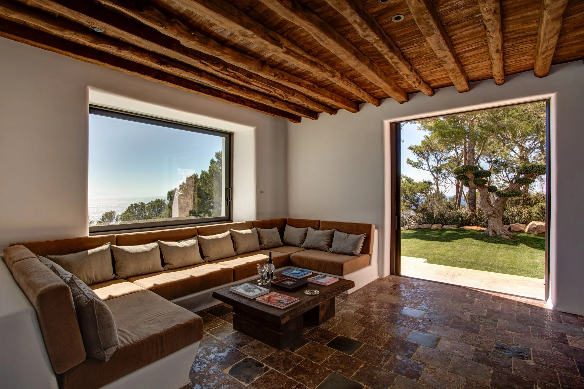 Living room with large windows and wooden beamed ceiling, outdoor view of trees and a lawn with a sculpted tree, tan sectional sofa with multiple pillows, coffee table with magazines, and tiled floor.