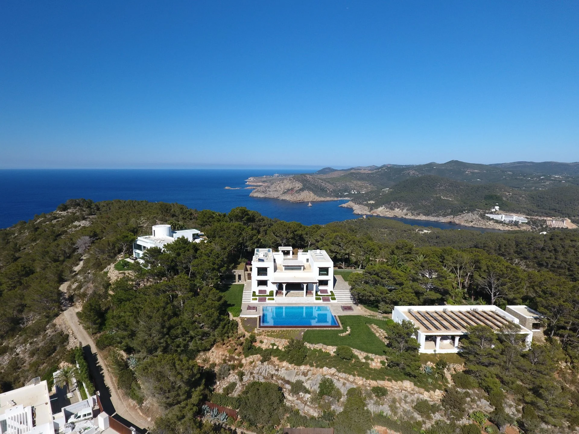 Aerial view of a coastal landscape with a modern white house featuring a large swimming pool, surrounded by lush green trees and hills, overlooking the ocean with a rocky coastline under a clear blue sky.