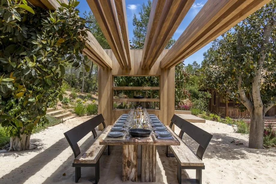 Outdoor dining area with a wooden table set with plates, glasses, and a black skillet, under a wooden pergola, surrounded by trees and greenery.