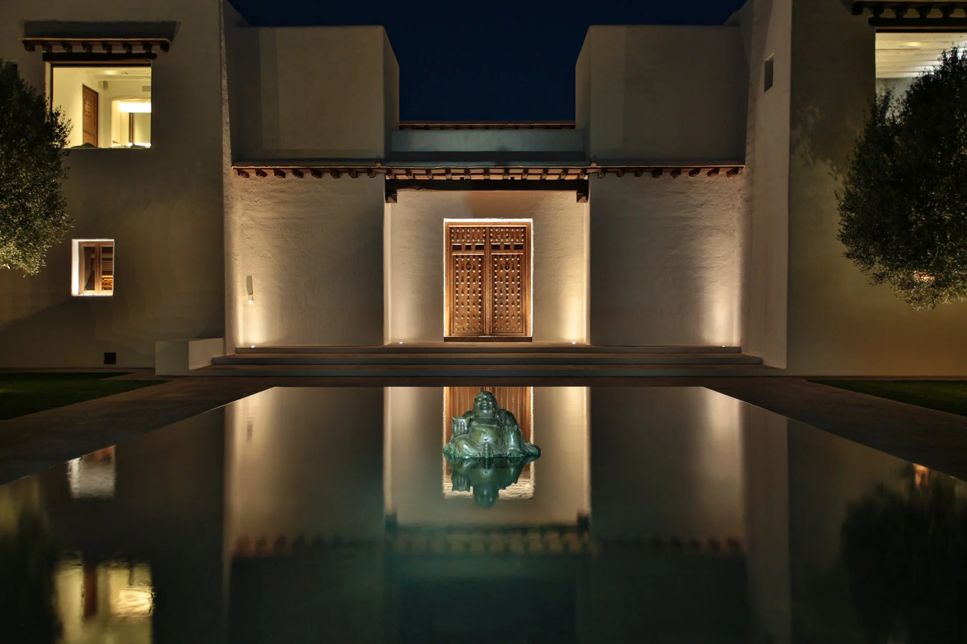 Night view of a modern building with white walls, wooden door, illuminated by exterior lights, flanked by trees, with a reflecting pool and a Buddha statue in the foreground.