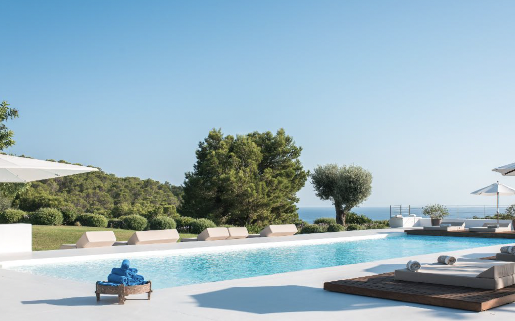 Luxury outdoor swimming pool area with lounge chairs, umbrellas, and a scenic view of greenery and the ocean in the background on a clear, sunny day.