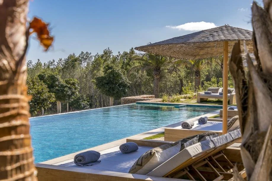 Swimming pool with lounge chairs and rolled towels under a thatched umbrella, surrounded by trees and clear blue sky.
