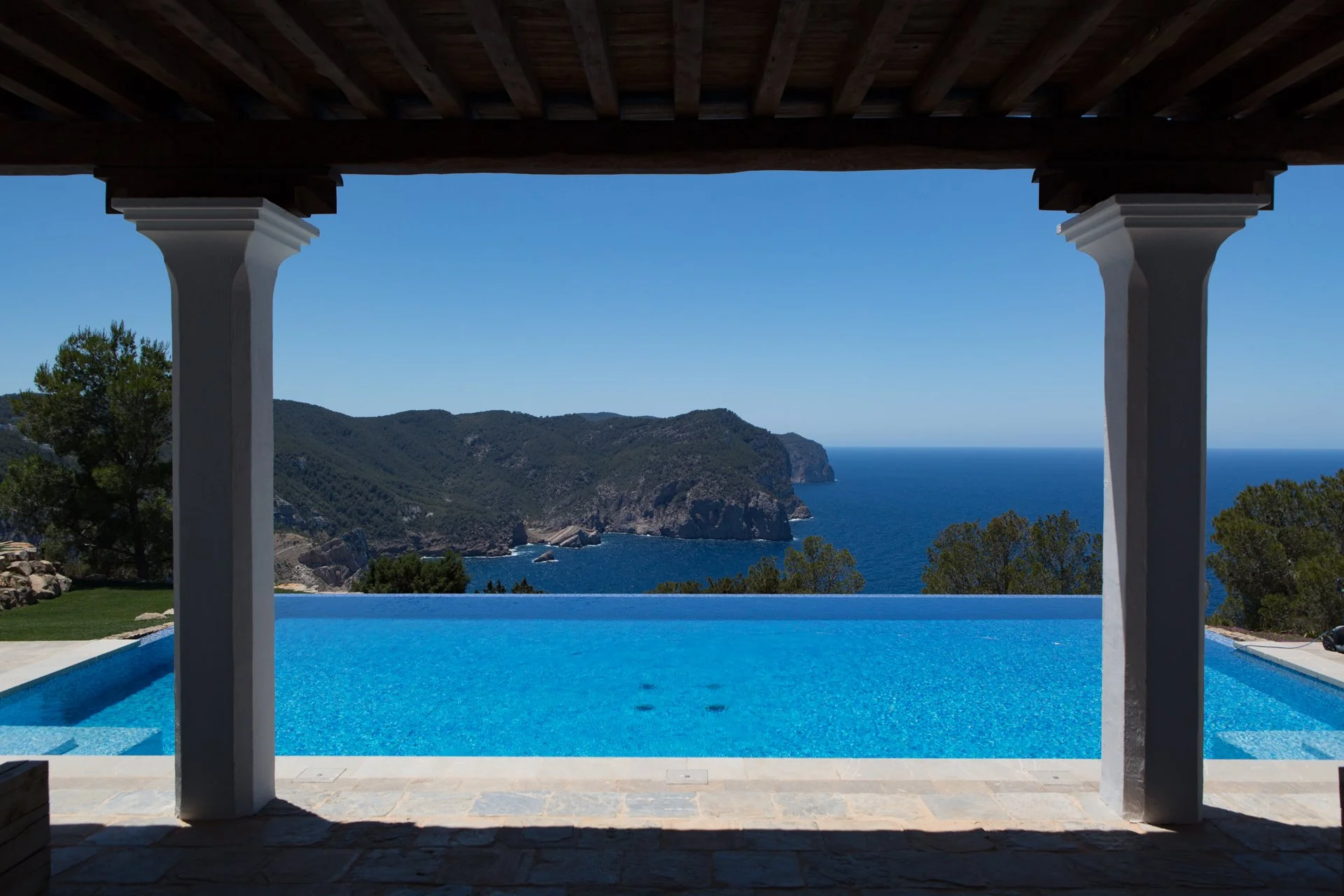 View of a blue infinity pool overlooking mountains and the ocean, framed by white pillars and a wooden ceiling.