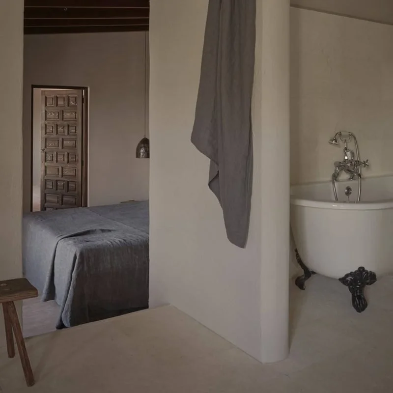 Interior of a bedroom with a bed, a hanging pendant light, a door with a geometric pattern, and a bathroom with a clawfoot bathtub and a gray towel hanging on a wall.