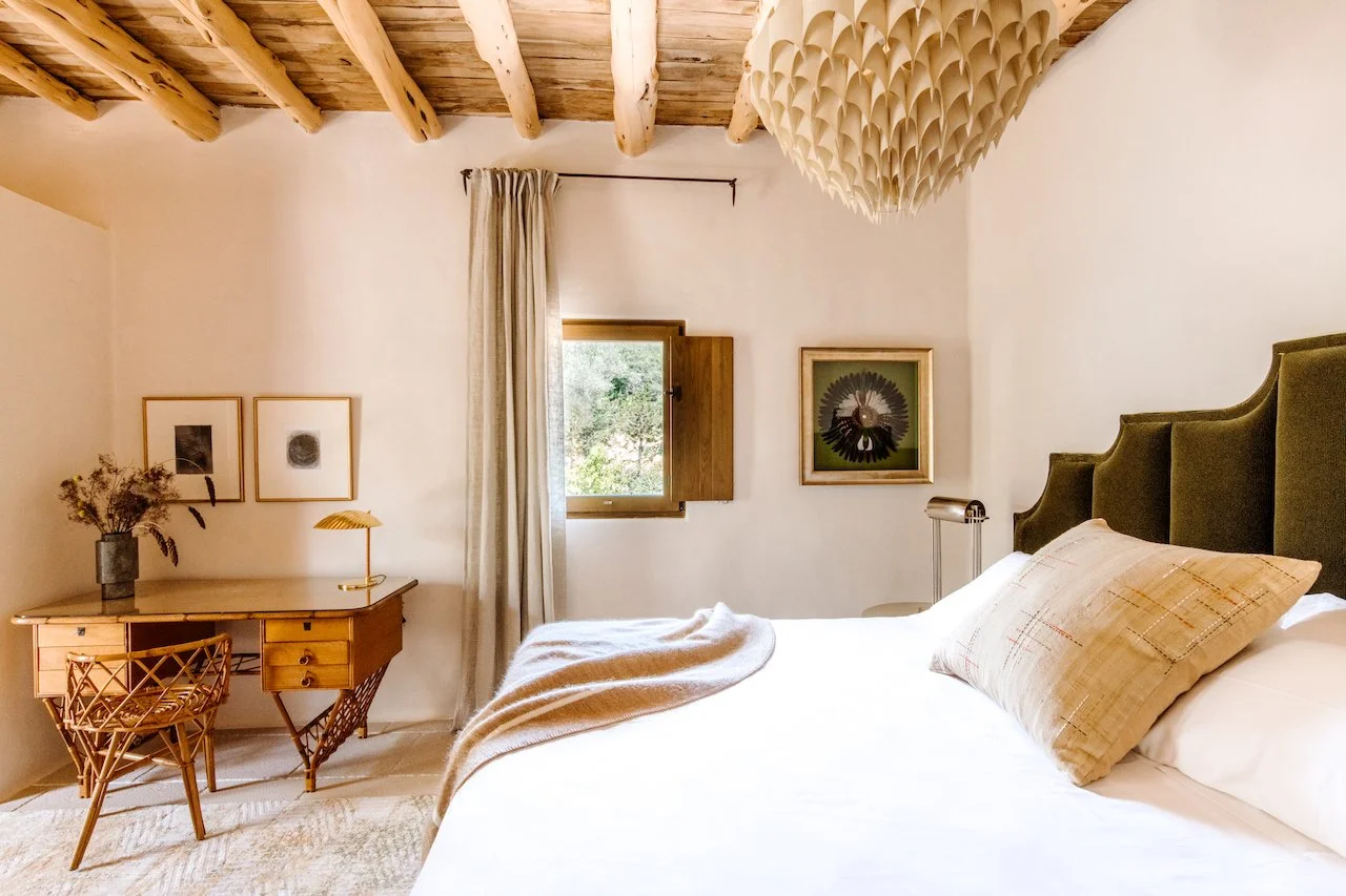 A cozy bedroom with a large bed featuring a padded green headboard, a beige pillow, and a cream-colored blanket, a wooden desk with a chair, framed artwork on the walls, a small window with curtains, and a wooden ceiling with exposed beams.