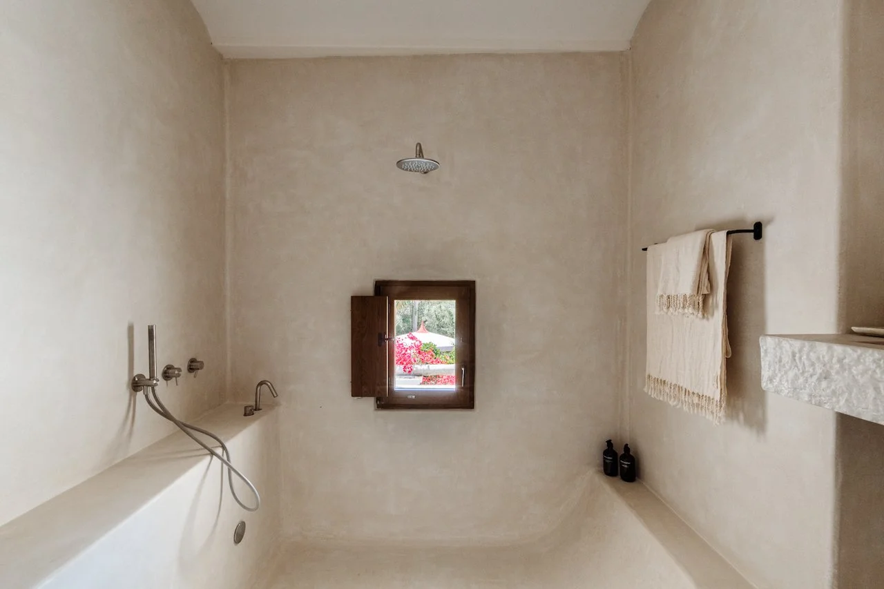 A minimalist bathroom with a small window showing a garden with pink flowers, a wall-mounted faucet, a handheld shower, a rolled towel on a black towel bar, and two black bottles in the corner.