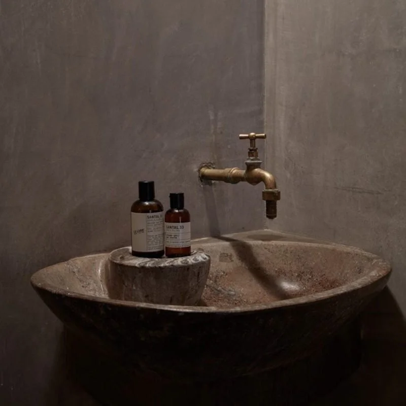 A rustic stone sink with a vintage brass faucet, two bottles of toiletries on a small stone platform, in a minimalistic bathroom with gray textured walls.