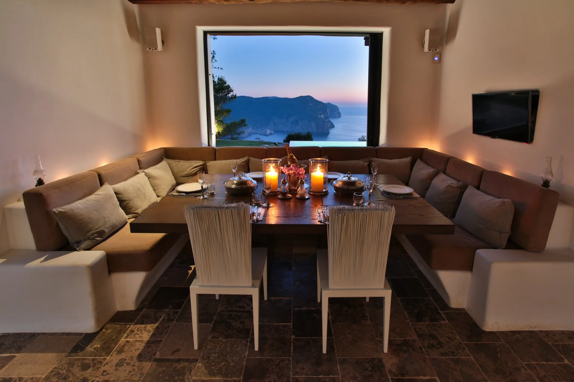 A dining room with a large wooden table set with plates, glasses, candles, and flowers. Cushioned chairs surround the table. There is a large window showing a scenic view of cliffs and the ocean at sunset. The room has a flat-screen TV on the wall an