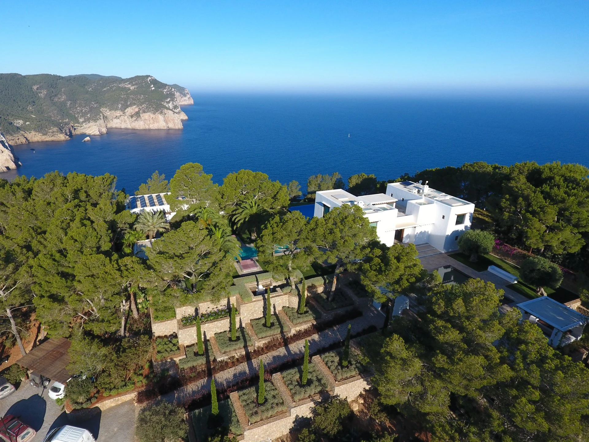 A scenic view of a coastal area with lush green trees, white modern buildings, a deep blue ocean with a sailboat, rocky cliffs, and clear sky.
