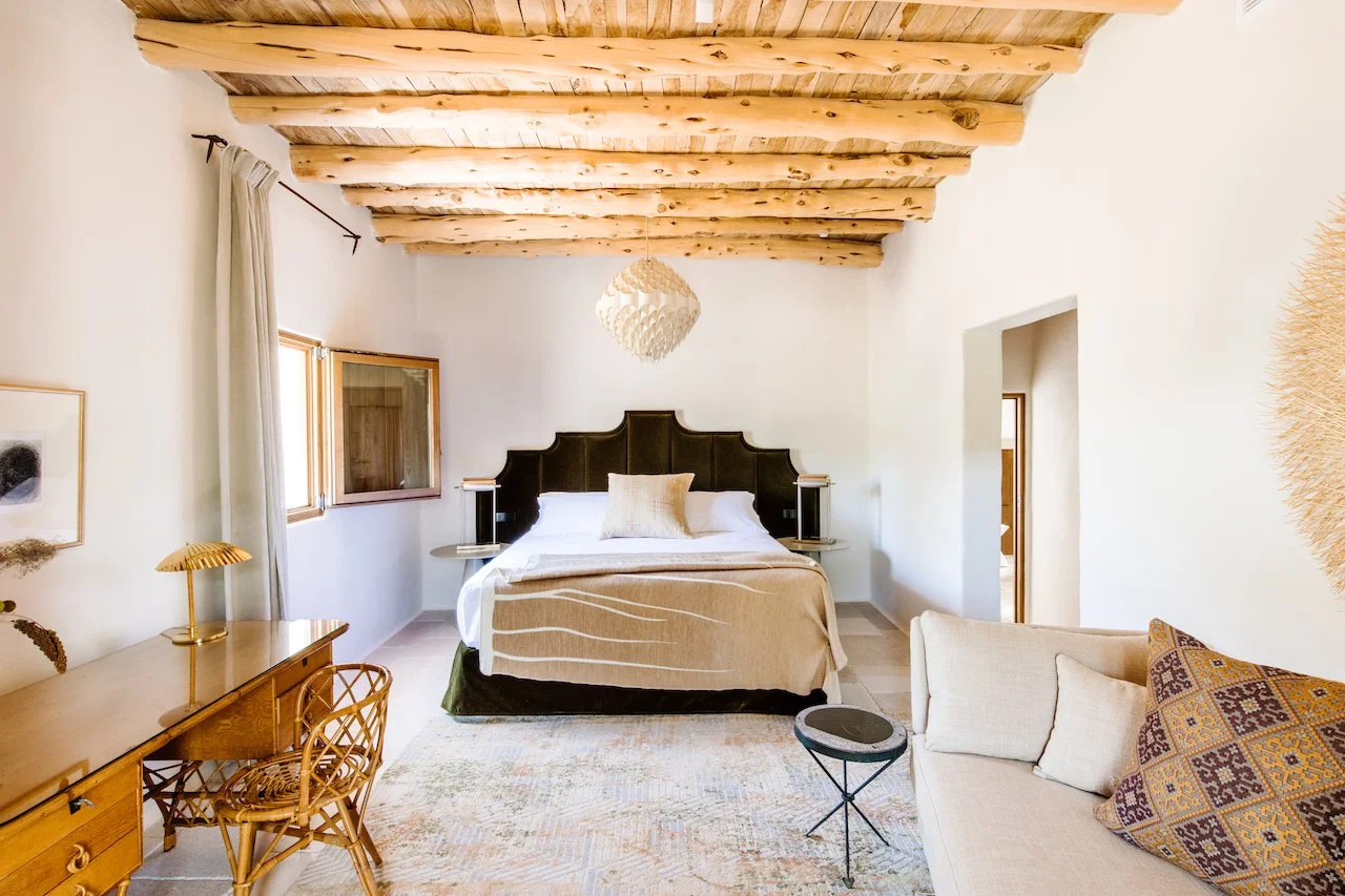 A bedroom with a wooden ceiling and white walls, featuring a large bed with a dark headboard, a beige blanket, and white pillows. There is a desk with a wicker chair and a gold lamp on the left, a window with white curtains, and a sofa with patterned