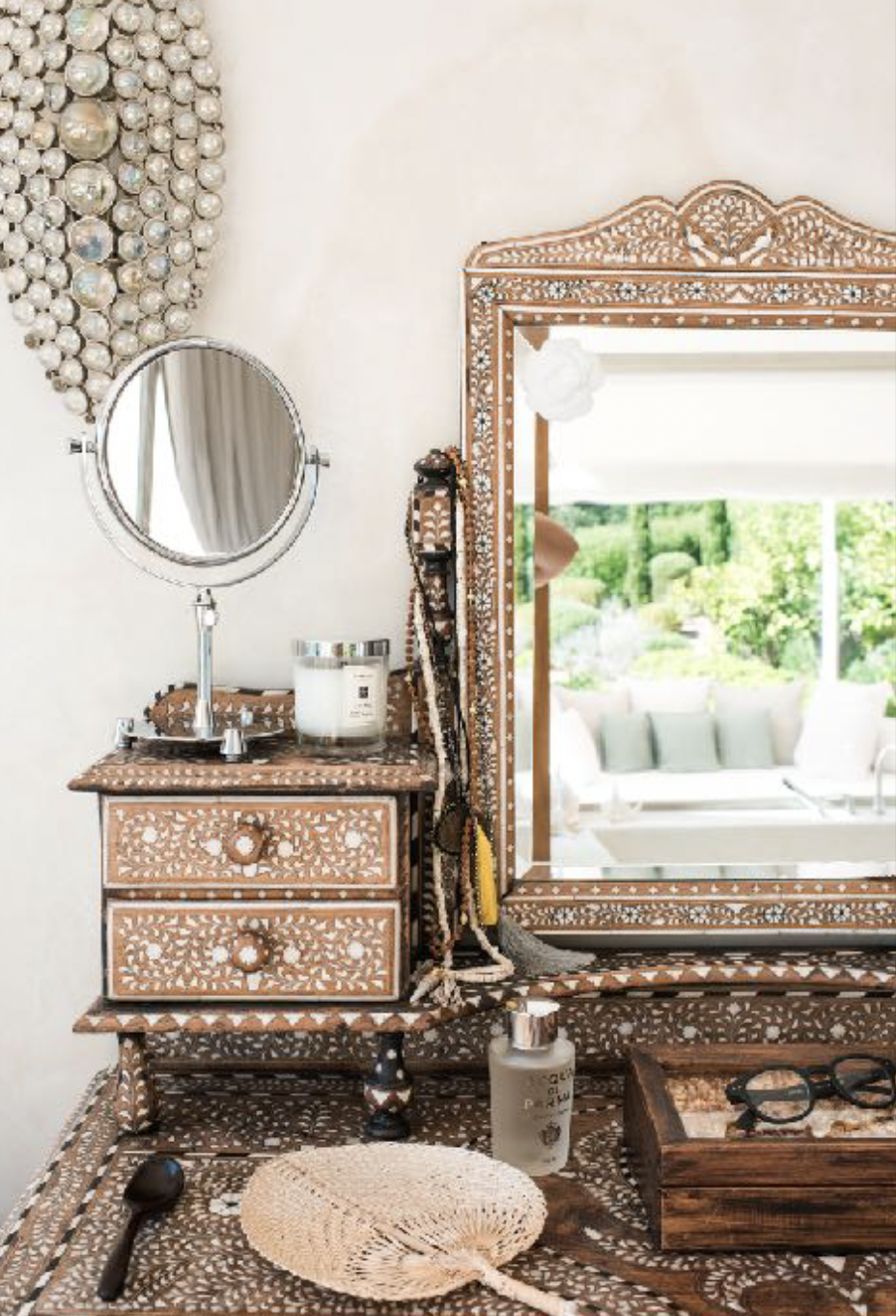 A vanity table with a mirror, jewelry, perfume, candles, sunglasses, a fan, and cleaning spray in a room with a window overlooking a garden.