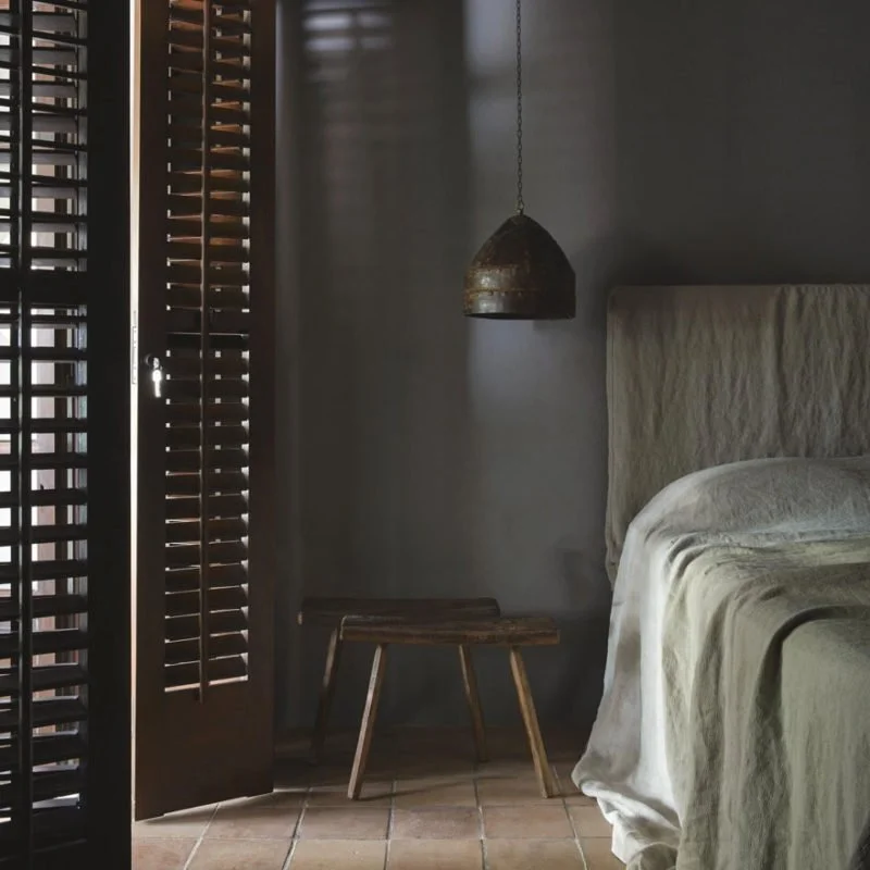 A bedroom corner with a wooden door with blinds, a small wooden stool, a hanging lamp, and part of a bed with beige linens.