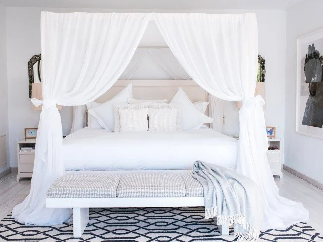White four-poster bed with sheer curtains, multiple pillows, a bench at the foot of the bed, a blanket draped over it, and side tables with lamps in a bright bedroom
