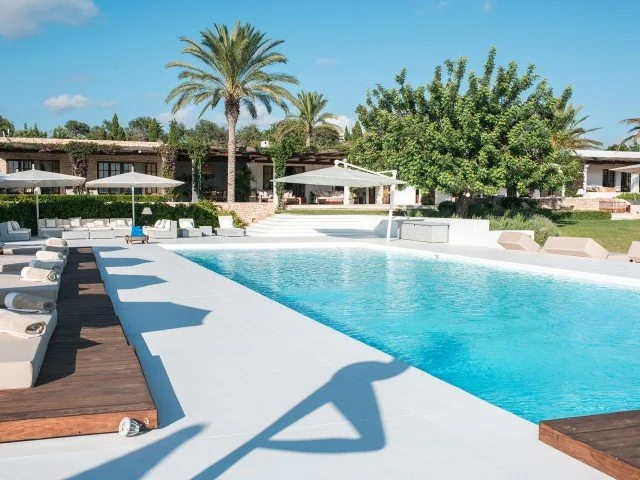 Luxury outdoor pool area at a resort with lounge chairs, umbrellas, and palm trees in the background on a sunny day.