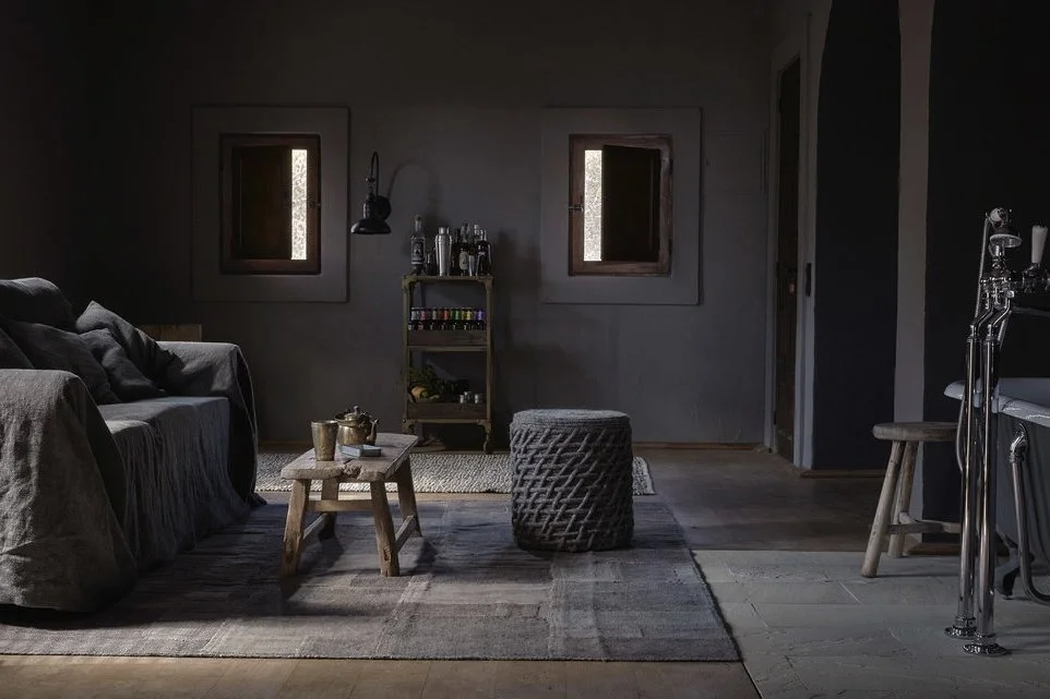 A dimly lit rustic living room with a dark sofa, a small wooden coffee table with teapots and cups, a woven ottoman, two small windows, a metal shelf with bottles and colorful spices, and a stone floor with a large rug.