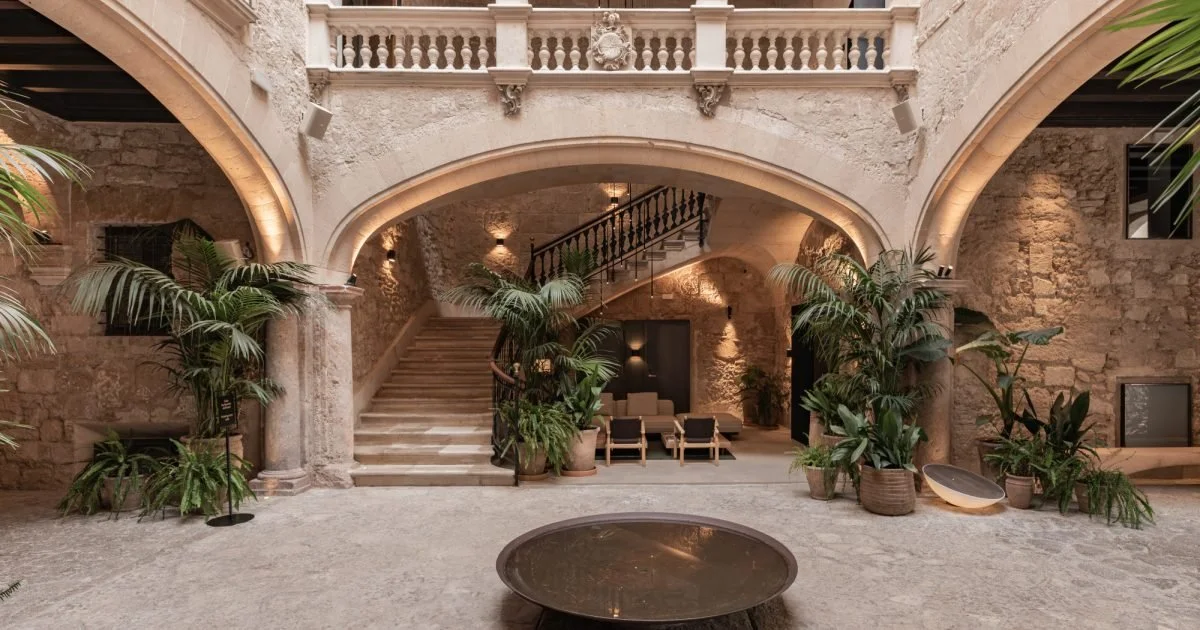 Interior of a historic stone building with a courtyard, featuring stone arches, a staircase, potted plants, and seating area with chairs and sofas.