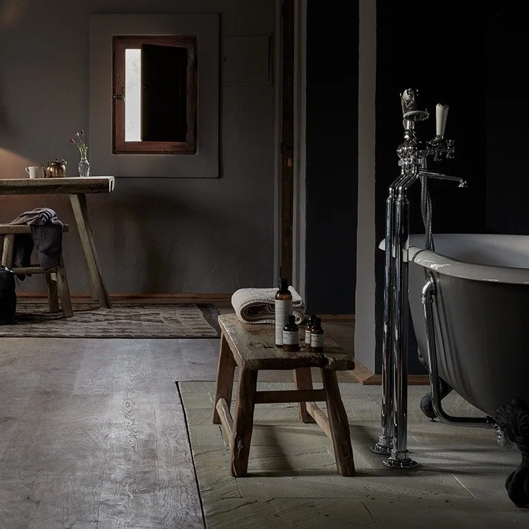 Rustic bathroom with a clawfoot bathtub, a wooden stool with bottles and towels, a dark wall, a small window, and a table with a flower in a vase.