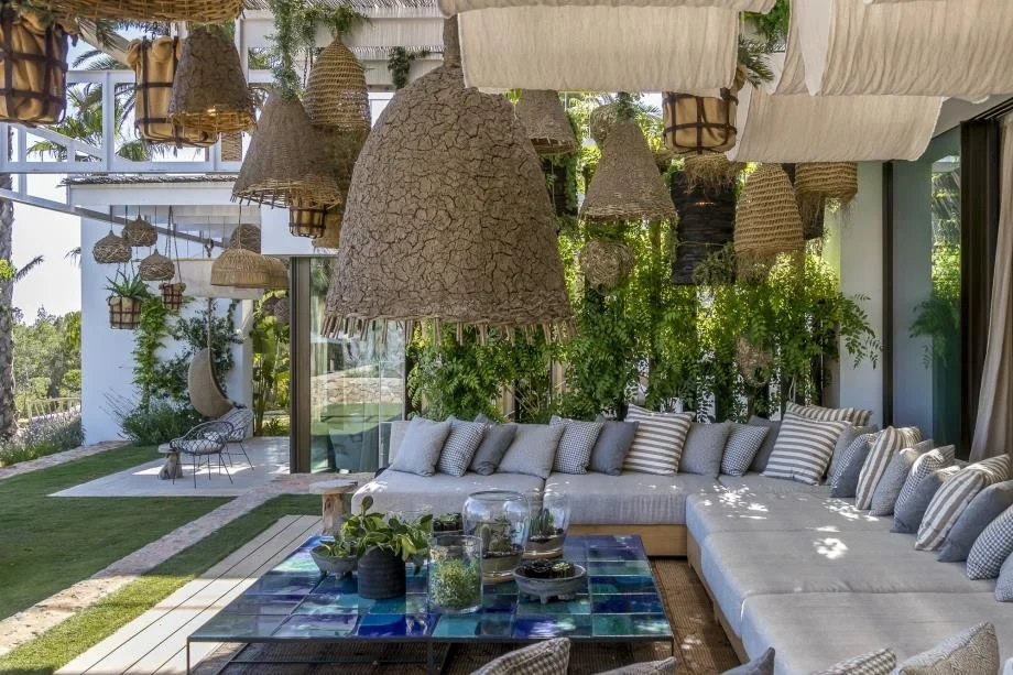Outdoor patio with a large gray sectional sofa with striped and solid throw pillows, a glass coffee table with decorative items, hanging wicker and fabric pendant lights, lush green plants, and a view of a garden with a white lounge chair in the back
