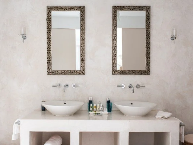 Double bathroom vanity with two vessel sinks, two framed mirrors, two wall-mounted light fixtures, and toiletries on the counter in a neutral-toned room.