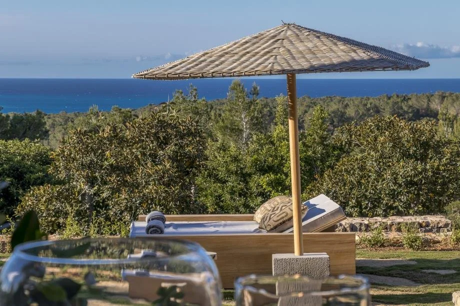 Beachside outdoor lounge area with a wooden lounge chair, a towel, a cushion, and a large straw umbrella overlooking lush greenery and the ocean in the background.